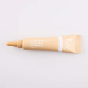 Das Bild zeigt eine beige Tube mit einer spitzen Dosierspitze, liegend auf einem beigen Hintergrund. Das Produkt gehört zur KRX Aesthetics-Reihe und trägt den Namen „All Day Glow Gel Cleanser“. Die Tube hat eine weiße Beschriftung mit einer dünnen weißen Linie und enthält 3,5 ml des Produkts. Die Beleuchtung ist sanft und gleichmäßig.