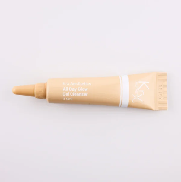 Das Bild zeigt eine beige Tube mit einer spitzen Dosierspitze, liegend auf einem beigen Hintergrund. Das Produkt gehört zur KRX Aesthetics-Reihe und trägt den Namen „All Day Glow Gel Cleanser“. Die Tube hat eine weiße Beschriftung mit einer dünnen weißen Linie und enthält 3,5 ml des Produkts. Die Beleuchtung ist sanft und gleichmäßig.