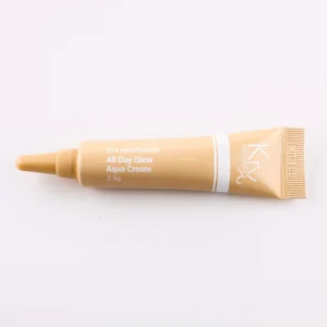 Das Bild zeigt eine weitere beige Tube mit einer spitzen Dosierspitze auf einem beigen Hintergrund. Das Produkt gehört zur KRX Aesthetics-Reihe und trägt den Namen „All Day Glow Aqua Cream“. Die Tube hat eine weiße Beschriftung mit einer dünnen weißen Linie und enthält 3,5 g. Die Beleuchtung ist sanft und gleichmäßig.