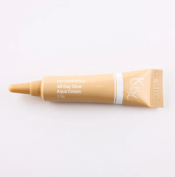 Das Bild zeigt eine weitere beige Tube mit einer spitzen Dosierspitze auf einem beigen Hintergrund. Das Produkt gehört zur KRX Aesthetics-Reihe und trägt den Namen „All Day Glow Aqua Cream“. Die Tube hat eine weiße Beschriftung mit einer dünnen weißen Linie und enthält 3,5 g. Die Beleuchtung ist sanft und gleichmäßig.