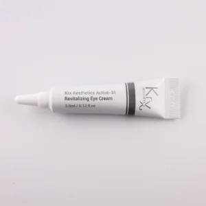 Das Bild zeigt eine kleine Tube mit einer spitzen Dosierspitze, liegend auf einem beigen Hintergrund. Das Produkt stammt von KRX Aesthetics und trägt den Namen „Active-31 Revitalizing Eye Cream“. Die Tube ist weiß mit einem schlichten schwarzen und grauen Designstreifen. Die Aufschrift lautet: „KRX Aesthetics Active-31 Revitalizing Eye Cream“, mit einer Füllmenge von 3.5 ml (0.12 FL.OZ.). Das Bild ist gleichmäßig beleuchtet, mit einem sanften Beige als Hintergrund.