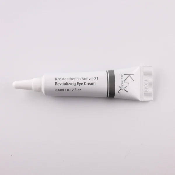 Das Bild zeigt eine kleine Tube mit einer spitzen Dosierspitze, liegend auf einem beigen Hintergrund. Das Produkt stammt von KRX Aesthetics und trägt den Namen „Active-31 Revitalizing Eye Cream“. Die Tube ist weiß mit einem schlichten schwarzen und grauen Designstreifen. Die Aufschrift lautet: „KRX Aesthetics Active-31 Revitalizing Eye Cream“, mit einer Füllmenge von 3.5 ml (0.12 FL.OZ.). Das Bild ist gleichmäßig beleuchtet, mit einem sanften Beige als Hintergrund.