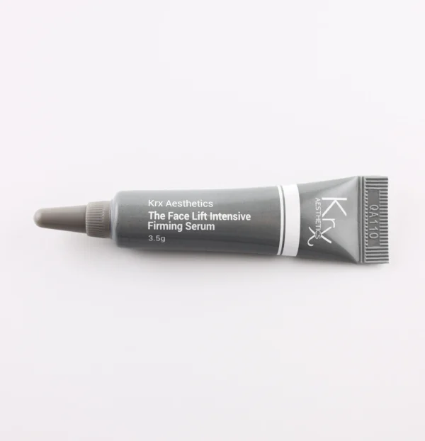 Das Bild zeigt eine ähnliche kleine Tube mit einer spitzen Dosierspitze, ebenfalls auf einem beigen Hintergrund. Das Produkt stammt von KRX Aesthetics und heißt „The Face Lift Intensive Firming Serum“. Die Tube ist schwarz-grau mit weißem Text und einer feinen weißen Linie. Die Füllmenge beträgt 3,5 g. Das Bild hat eine gleichmäßige, sanfte Beleuchtung.