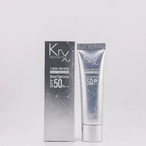 Das Bild zeigt ein Kosmetikprodukt der Marke „KrX Aesthetics“ in einer silbernen Tube. Die Tube liegt schräg auf einer glatten, hellgrauen Oberfläche. Auf der Vorderseite der Tube ist in heller Schrift unter anderem der Hinweis „SPF 50+“ (ein hoher Sonnenschutzfaktor) zu erkennen. Links neben der Tube befinden sich zwei nebeneinander aufgetragene Produktproben in cremiger Konsistenz. Die Farbe ist ein warmes Beige mit einem leichten Orangeton. Beide Proben haben eine tropfenartige, nach oben spitz zulaufende Form und glänzen leicht, was auf eine feuchtigkeitsspendende oder pflegende Textur hindeutet. Im Hintergrund ist der Rand eines Laptop-Bildschirms zu sehen, auf dem das Bild offenbar angezeigt wird. Darüber hinaus sind unscharf einige Pflanzen und Blüten zu erkennen.