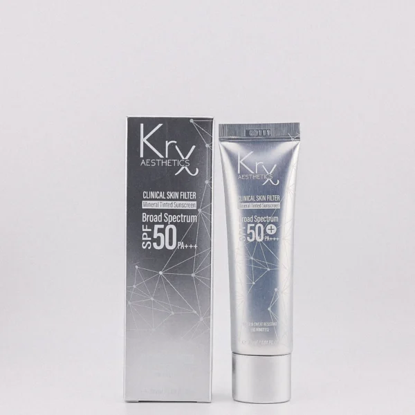 Das Bild zeigt ein Kosmetikprodukt der Marke „KrX Aesthetics“ in einer silbernen Tube. Die Tube liegt schräg auf einer glatten, hellgrauen Oberfläche. Auf der Vorderseite der Tube ist in heller Schrift unter anderem der Hinweis „SPF 50+“ (ein hoher Sonnenschutzfaktor) zu erkennen. Links neben der Tube befinden sich zwei nebeneinander aufgetragene Produktproben in cremiger Konsistenz. Die Farbe ist ein warmes Beige mit einem leichten Orangeton. Beide Proben haben eine tropfenartige, nach oben spitz zulaufende Form und glänzen leicht, was auf eine feuchtigkeitsspendende oder pflegende Textur hindeutet. Im Hintergrund ist der Rand eines Laptop-Bildschirms zu sehen, auf dem das Bild offenbar angezeigt wird. Darüber hinaus sind unscharf einige Pflanzen und Blüten zu erkennen.