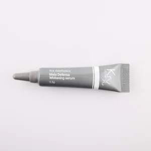 Dieses Bild zeigt eine weitere kleine Tube von KRX Aesthetics, die den Namen „Mela Defense Whitening Serum“ trägt. Die Tube ist schwarz-grau, mit weißem Schriftzug und einer dünnen weißen Linie. Die Füllmenge beträgt 3,5 g. Das Produkt liegt auf einem beigen Hintergrund, und die Beleuchtung ist weich und gleichmäßig.