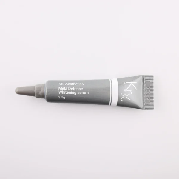 Dieses Bild zeigt eine weitere kleine Tube von KRX Aesthetics, die den Namen „Mela Defense Whitening Serum“ trägt. Die Tube ist schwarz-grau, mit weißem Schriftzug und einer dünnen weißen Linie. Die Füllmenge beträgt 3,5 g. Das Produkt liegt auf einem beigen Hintergrund, und die Beleuchtung ist weich und gleichmäßig.