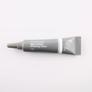 Das Bild zeigt eine kleine, dunkle Tube mit einer spitzen Dosierspitze, liegend auf einem beigen Hintergrund. Das Produkt gehört zur Marke KRX Aesthetics und trägt den Namen „Mela Defense Whitening Cream“. Die Tube ist schwarz-grau mit einem weißen Schriftzug und einer dünnen weißen Linie. Der Inhalt beträgt 3,5 g. Die Beleuchtung ist weich und gleichmäßig, was das Produkt gut erkennbar macht.