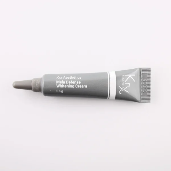 Das Bild zeigt eine kleine, dunkle Tube mit einer spitzen Dosierspitze, liegend auf einem beigen Hintergrund. Das Produkt gehört zur Marke KRX Aesthetics und trägt den Namen „Mela Defense Whitening Cream“. Die Tube ist schwarz-grau mit einem weißen Schriftzug und einer dünnen weißen Linie. Der Inhalt beträgt 3,5 g. Die Beleuchtung ist weich und gleichmäßig, was das Produkt gut erkennbar macht.