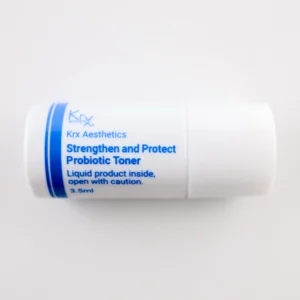 Hier ist ein ähnlicher kleiner, weißer Zylinderbehälter zu sehen, diesmal mit blauer Beschriftung. Das Produkt gehört ebenfalls zur KRX Aesthetics-Reihe und trägt den Namen „Strengthen and Protect Probiotic Toner“. Der Behälter enthält 3,5 ml Flüssigkeit und trägt ebenfalls die Warnung: „Liquid product inside, open with caution.“ Der Hintergrund ist beige, und die Beleuchtung ist sanft und gleichmäßig.