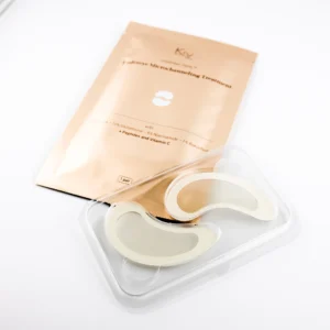 Das Bild zeigt eine Packung mit Augenpads der Marke Krx Aesthetics, die für ein mikroverkapseltes Hautpflege-Treatment entwickelt wurden. In der Mitte des Bildes liegt ein beigefarbenes Sachet mit dem Aufdruck „Eyebrow Microchanneling Treatment“. Darunter sind Wirkstoffe wie 5 % Glutathion, 3 % Niacinamide, 1 % Bakuchiol, Peptide und Vitamin C aufgeführt. Im Vordergrund ist eine transparente Kunststoffschale zu sehen, in der sich ein Paar gebogener Augenpads befindet – hautfarben, leicht transparent und mit einem leichten Glanz, was auf eine feuchtigkeitsspendende oder aktive Schicht hindeutet. Die Pads haben eine ergonomische Form, die sich der Augenpartie anpasst. Auf der Verpackung ist angegeben: „1 pair“.