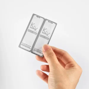 Das Bild zeigt eine Hand, die zwei kleine silberfarbene Sachets mit weißer Beschriftung in die Kamera hält. Es handelt sich um die “KrX Aesthetics Repair Cream”, eine Creme in Probiergröße, verpackt in Einzeldosen (Sachets). Die Verpackung ist schlicht und professionell gestaltet. Auf den Sachets steht, dass die Creme speziell für die Pflege nach Hautbehandlungen wie MTS (Microneedling), Laser oder Peels gedacht ist.