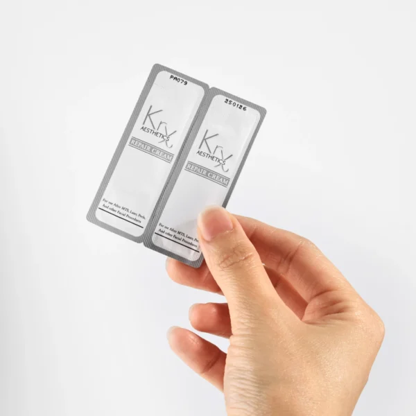 Das Bild zeigt eine Hand, die zwei kleine silberfarbene Sachets mit weißer Beschriftung in die Kamera hält. Es handelt sich um die “KrX Aesthetics Repair Cream”, eine Creme in Probiergröße, verpackt in Einzeldosen (Sachets). Die Verpackung ist schlicht und professionell gestaltet. Auf den Sachets steht, dass die Creme speziell für die Pflege nach Hautbehandlungen wie MTS (Microneedling), Laser oder Peels gedacht ist.