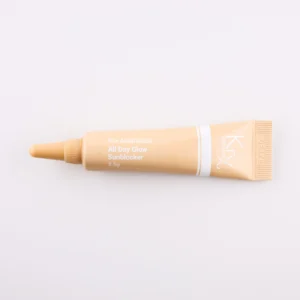 Hier ist eine weitere beige Tube mit einer spitzen Dosierspitze zu sehen. Das Produkt gehört zur KRX Aesthetics-Reihe und trägt den Namen „All Day Glow Sunblocker“. Die Tube hat eine weiße Beschriftung und enthält 3,5 g des Produkts. Das Produkt liegt auf einem beigen Hintergrund mit gleichmäßiger Beleuchtung.