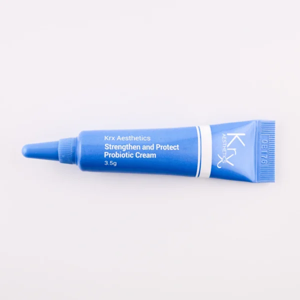 Hier ist eine weitere blaue Tube mit ähnlichem Design zu sehen. Sie gehört ebenfalls zur KRX Aesthetics-Reihe und trägt den Namen „Strengthen and Protect Probiotic Cream“. Die Tube hat eine weiße Beschriftung mit einer dünnen weißen Linie und enthält 3,5 g des Produkts. Das Produkt liegt auf einem beigen Hintergrund mit gleichmäßiger Beleuchtung.